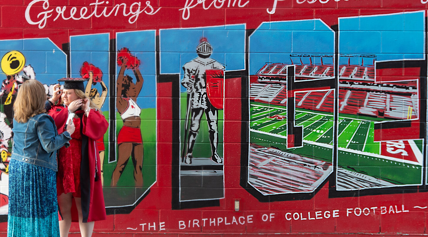 Parent helping student get ready in Commencement regalia in front of a painted Rutgers football wall mural